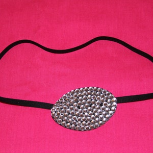 May include: A black elasticated headband featuring an oval eye patch. The eye patch is embellished with numerous small, shiny, silver rhinestones. The background is a vibrant, solid pink.