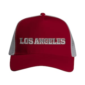 May include: A red trucker hat with the embroidered text "LOS ANGELES" in silver. The hat features a mesh back in a light gray color and a curved brim.
