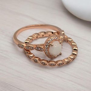 Può includere: Set di anelli di fidanzamento in oro rosa. Il set include un anello con una pietra bianca circondata da piccole pietre trasparenti e una fascia abbinata con piccole pietre trasparenti. Gli anelli sono su una superficie a venature di legno chiaro.