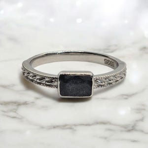 Op de afbeelding: Een zilveren ring met een rechthoekige, donkergrijze steen in het midden. De band heeft een gedetailleerd, gevlochten ontwerp. De ring is geplaatst tegen een witte marmeren achtergrond. De ring is gemarkeerd met "925".