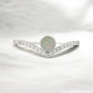 May include: A silver ring with a chevron design. The ring features a round, iridescent opal stone at the centre and is accented with small, clear gemstones along the chevron band. The ring is set against a white background.