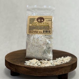 May include: A clear plastic bag of "Beef Stew & Pot Roast Seasoning" sits on a small, round, wooden stand. The seasoning is a mix of white, green, and brown herbs and spices. The stand has a dark brown finish and three small legs.