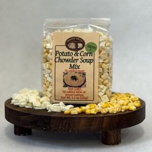 May include: A clear bag of Muddy Trail Potato & Corn Chowder Soup Mix, with the text "SERVES 8". The bag is filled with dried corn and potato pieces. The product is displayed on a round, wooden tray. The net weight is 5.5 oz (155g).