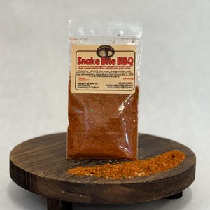May include: A clear plastic packet of "Snake Bite BBQ" seasoning sits on a small, round, dark wooden stand. The packet has a brown label with text and a logo. A small pile of seasoning is next to the packet. The seasoning appears to be a reddish-brown color.