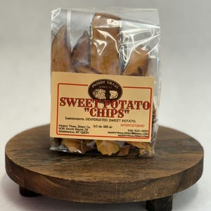 May include: A clear plastic bag filled with sweet potato chips, with a label that reads "Sweet Potato Chips." The bag is sitting on a round, dark wooden serving board with three small legs.