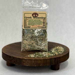 May include: A clear plastic bag of Greek seasoning sits on a small, round, dark wooden tray. The label reads "Greek Seasoning" with a list of ingredients and instructions. Some seasoning is scattered on the tray.