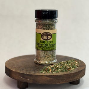 May include: A clear spice jar with a black lid, labeled "Salt Free Olive Oil Bread Dipping Seasoning" sits on a small wooden stand. A small pile of seasoning is next to the jar. The label is green and white.