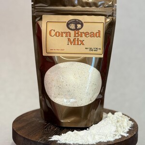 May include: A brown package of "Corn Bread Mix" on a small wooden stand. The package has a clear window showing the mix inside. A small pile of white powder sits on the stand next to the package. The package is labeled "Muddy Trail".
