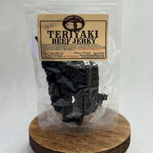 May include: A clear plastic bag of Teriyaki Beef Jerky sits on a small wooden stand. The jerky is dark brown and appears to be dried and sliced. The bag has a label with the text "Teriyaki Beef Jerky" and the Muddy Trail Jerky Co. logo.