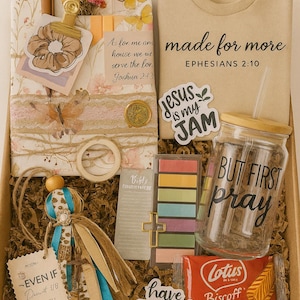 Puede incluir: Una caja de regalo llena de varios artículos, incluyendo una camiseta beige con el texto "made for more", un frasco de vidrio con pajita y una pegatina "Jesus is my Jam". Otros artículos incluyen un marcapáginas, una muñeca y una galleta.