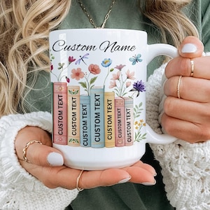 以下が含まれることがあります： カラフルな花と蝶のフローラルデザインが施された白いセラミックマグ。マグカップには「CUSTOM TEXT」の文字が入った本が並んでおり、「Custom Name」の文字も入っています。読書好きの方へのプレゼントに最適です。