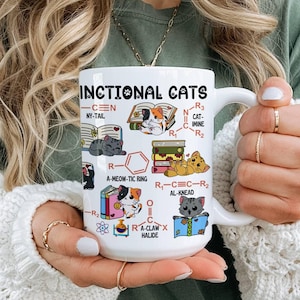 Könnte beinhalten: Weiße Keramik-Tasse mit der Aufschrift "INCTIONAL CATS" und Illustrationen von Katzen und chemischen Strukturen. Die Tasse hat einen Henkel und wird von einer Person gehalten, die einen weißen Pullover und ein grünes T-Shirt trägt.