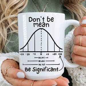 Op de afbeelding: Witte keramische mok met de tekst "Don't be mean, Be Significant" en een klokvormig diagram. De mok heeft een grote handgreep. De mok wordt vastgehouden door een persoon die een groen shirt en een witte trui draagt.