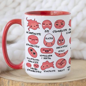Pode incluir: Caneca de cerâmica branca com interior e alça vermelhos, com ilustrações de desenhos animados de tipos de células sanguíneas como Acanthocyte e Sickle. A caneca está sobre uma superfície de madeira contra um fundo branco macio.