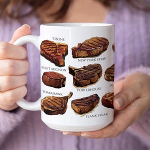 May include: White ceramic mug featuring illustrations of various steak cuts, including T-bone, New York strip, and filet mignon. The mug has a large handle and is held by a person wearing a purple sweater.