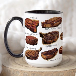 May include: White ceramic mug with a black handle and rim, featuring illustrations of various steak cuts, including T-bone, New York strip, filet mignon, and porterhouse. The mug is displayed on a wooden surface.