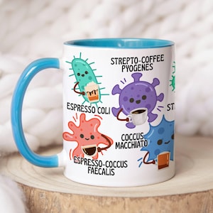 Könnte beinhalten: Weiße Keramik-Tasse mit blauem Henkel und Innenraum. Die Tasse zeigt Cartoon-Illustrationen von Mikroben, die jeweils eine Kaffeetasse halten, mit Textetiketten wie "Espresso Coli" und "Strepto-Coffee Pyogenes."