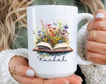 May include: White ceramic mug featuring a watercolor illustration of an open book with colorful wildflowers. The name "Rachel" is written in script below the book. The mug has a large handle and is held by a person.
