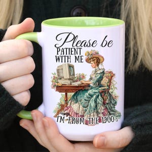 May include: White ceramic mug with a green interior and handle. The mug features a vintage illustration of a woman in a Victorian dress using a computer, with the text "Please be patient with me, I'm from the 1900s."