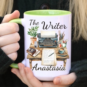 May include: White ceramic mug with a green interior and handle. The mug features a detailed illustration of a writer's desk with a typewriter, books, and the text "The Writer Anastasia."