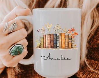 May include: White ceramic mug featuring a watercolor illustration of a stack of books with wildflowers. The name "Amelia" is written in cursive below the books. The mug is held by a person.