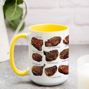 May include: White ceramic mug with a yellow interior and handle. The mug features illustrations of various cuts of steak, including T-bone, filet mignon, and New York strip. The mug is a kitchen and dining accessory.