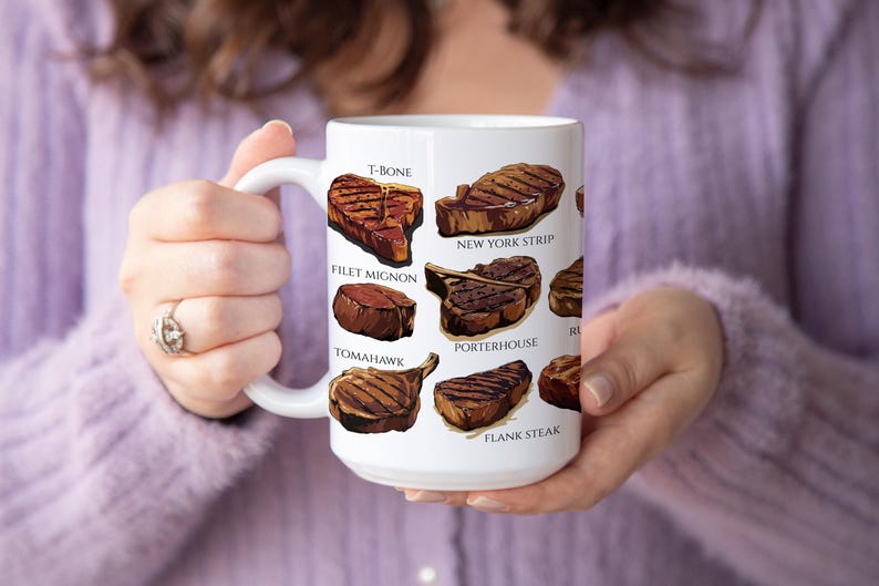 May include: White ceramic mug featuring illustrations of various steak cuts, including T-bone, New York strip, and filet mignon. The mug has a large handle and is held by a person wearing a purple sweater.