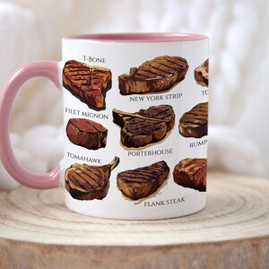 May include: White ceramic mug with a pink handle and rim, featuring illustrations of various steak cuts like T-bone, New York strip, and filet mignon. The mug is displayed on a wooden surface.