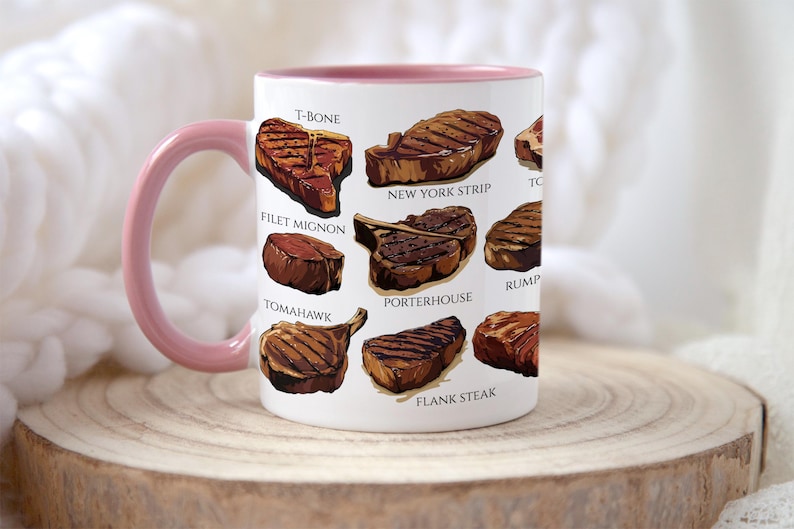 May include: White ceramic mug with a pink handle and rim, featuring illustrations of various steak cuts like T-bone, New York strip, and filet mignon. The mug is displayed on a wooden surface.