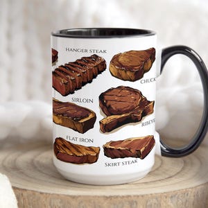 May include: A white ceramic mug with a black handle and rim, featuring illustrations of various cuts of steak, including hanger steak, sirloin, and ribeye. The mug is displayed on a wooden surface.