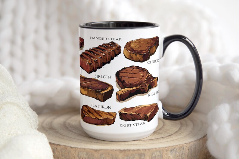 May include: A white ceramic mug with a black handle and rim, featuring illustrations of various cuts of steak, including hanger steak, sirloin, and ribeye. The mug is displayed on a wooden surface.