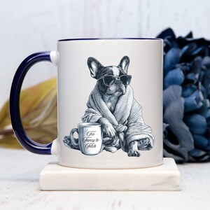 May include: White ceramic mug with a navy blue handle and rim. The mug features a black and white illustration of a French Bulldog wearing sunglasses and a robe, holding a mug that says "Too Fancy to Fetch."