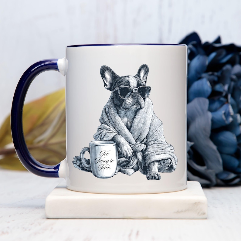 May include: White ceramic mug with a navy blue handle and rim. The mug features a black and white illustration of a French Bulldog wearing sunglasses and a robe, holding a mug that says "Too Fancy to Fetch."
