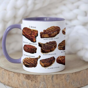 May include: White ceramic mug with a purple interior and handle, featuring illustrations of various steak cuts like T-bone, filet mignon, and New York strip. The mug is set on a wooden surface against a soft, white background.