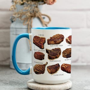 May include: White ceramic mug with a blue handle and interior, featuring illustrations of various steak cuts like T-bone, filet mignon, and New York strip. The mug sits on a round, light-colored coaster.