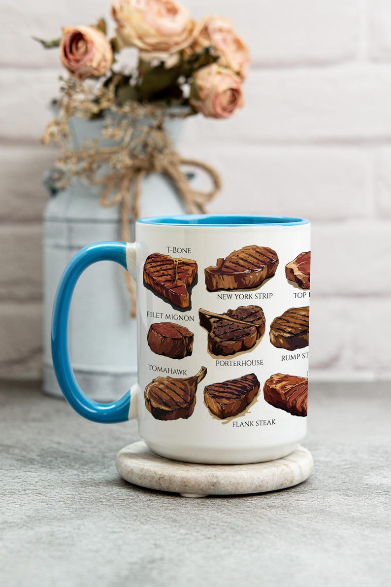 May include: White ceramic mug with a blue handle and interior, featuring illustrations of various steak cuts like T-bone, filet mignon, and New York strip. The mug sits on a round, light-colored coaster.