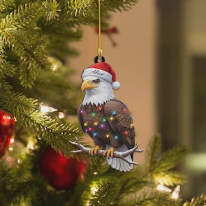 May include: A Christmas tree ornament featuring a bald eagle wearing a Santa hat with the name "Alice" and a string of colorful lights. The eagle is perched on a branch, with a gold string for hanging.