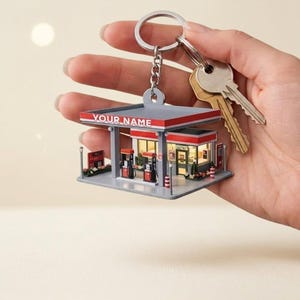 May include: A miniature gas station keychain with a red and gray canopy, red gas pumps, and the text "YOUR NAME". The keychain is attached to a silver keyring with two keys.