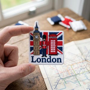 May include: A square embroidered patch featuring the Union Jack flag as a background. The design includes a tan Big Ben clock tower, a British guard in a red uniform, and a red telephone booth with the word TELEPHONE. The word London is in blue.
