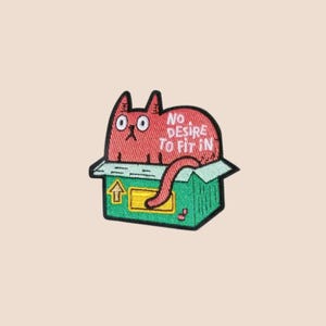 May include: Embroidered patch featuring a red cat inside a green box. The cat has large, round eyes and the text "NO DESIRE TO FIT IN" is above it. The box has a yellow arrow symbol.