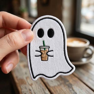 May include: A white embroidered patch shaped like a ghost with black stitched outlines and eyes. The ghost figure is holding a small tan-colored iced coffee cup with a green straw, featuring detailed brown stitching for the beverage and ice cubes.