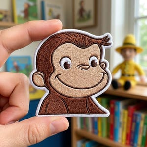 May include: Embroidered patch featuring a smiling cartoon monkey with brown fur and a white border. The monkey has large eyes and a cheerful expression. The patch is held up by a hand, with a blurred background of books and a toy.