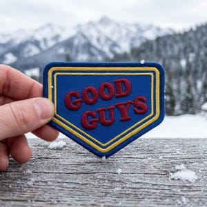 May include: A blue patch with the words "GOOD GUYS" in red, outlined in yellow. The patch has a shield shape and is held against a snowy mountain backdrop.