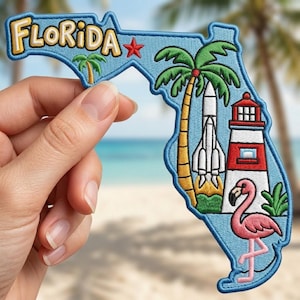 May include: Embroidered patch in the shape of Florida, featuring the word "FLORIDA" in yellow and blue. The patch includes images of a palm tree, rocket, lighthouse, and a pink flamingo. The patch is held against a blurred beach background.