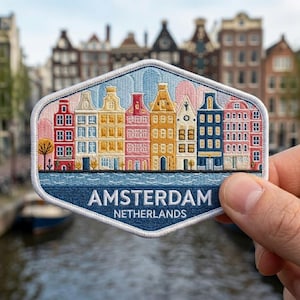May include: A hexagonal embroidered patch featuring a row of colorful canal houses in red, yellow, pink, and blue. Below the buildings is a blue water section with the white text AMSTERDAM NETHERLANDS. The patch has a white stitched border.