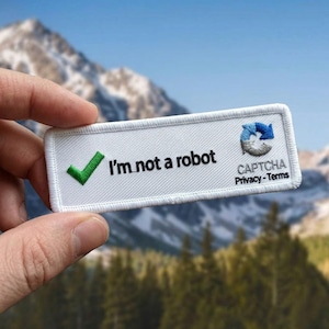 May include: A rectangular white patch with the text "I'm not a robot" and a green checkmark. The patch also features a CAPTCHA logo and the words "Privacy - Terms" in black. The background shows a mountain range and trees.