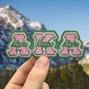 May include: Embroidered patch featuring the Greek letters Alpha Kappa Alpha in pink with a green border. The words "Alpha" and "Kappa" are stitched in green. The patch is held in front of a mountain backdrop.