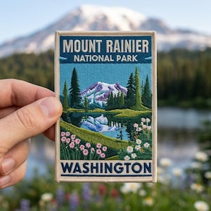 May include: A rectangular embroidered patch featuring a landscape of Mount Rainier National Park in Washington. The design includes a snow-capped mountain, evergreen trees, a blue lake, and pink and white wildflowers with text at the top and bottom.