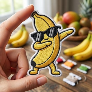 May include: Embroidered patch featuring a yellow banana wearing black sunglasses, in a dabbing pose. The patch has a white border and is held up in front of a wooden surface with fruit in the background. The patch is a fun, playful accessory.