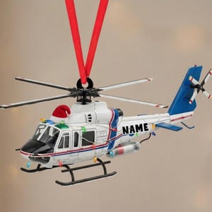 May include: A white and blue helicopter Christmas ornament with a red Santa hat and string lights. The helicopter has a black base and tail, with the word "NAME" on the side. It hangs from a red ribbon.